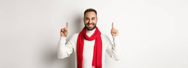 Vánoční nákupy a zimní prázdniny koncept. Usmívající se muž s vousy ukazující logo, ukazující prsty nahoru a vypadá šťastně, stojí v červené šále se svetrem, bílé pozadí. - Fotografie, Obrázek