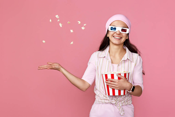 smiling beautiful woman in 3d-glasses holding bucket with flying popcorn isolated on pink background - Zdjęcie, obraz