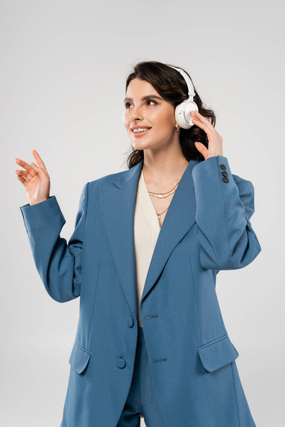 mujer morena sonriente en blazer azul y auriculares inalámbricos escuchando música y mirando hacia otro lado aislado en gris - Foto, Imagen