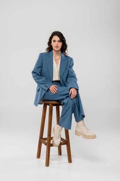 plná délka brunetky žena v modrém obleku a bílé boty sedí na dřevěné stoličce na šedém pozadí - Fotografie, Obrázek