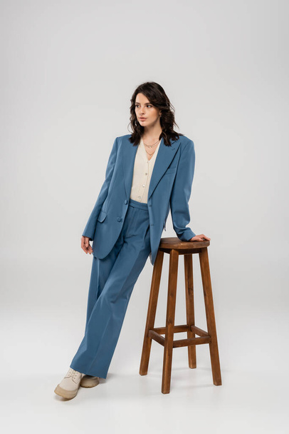 full length of brunette woman in blue suit posing near wooden stool on grey background - Photo, image