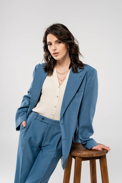 trendy woman in white jumper and blue suit posing with hand in pocket near wooden stool isolated on grey - Foto, imagen