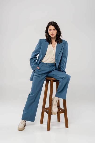 full length of young woman in blue and stylish suit holding hand in pocket while posing on stool on grey background - Фото, изображение