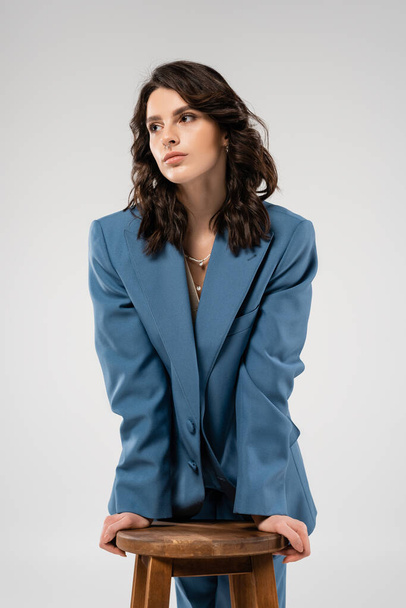 young and stylish woman in blue jacket looking away near wooden stool isolated on grey - Foto, Bild