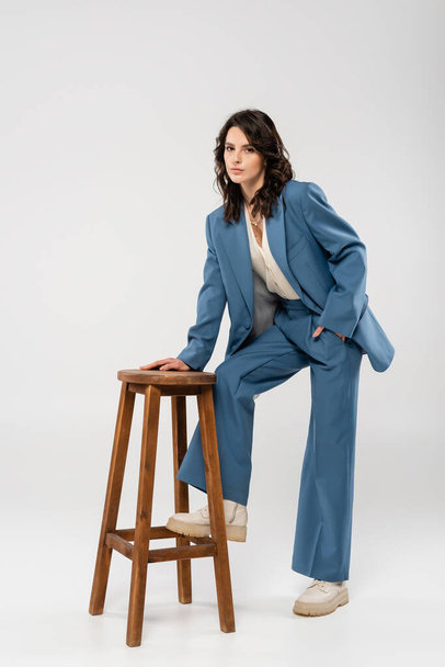 full length of fashionable woman in blue suit and boots posing with hand in pocket near wooden stool on grey background - Photo, Image