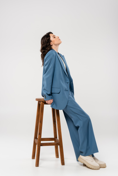 Seitenansicht einer brünetten Frau in blauem Anzug und Stiefeln, die auf einem Holzhocker auf grauem Hintergrund lehnt - Foto, Bild