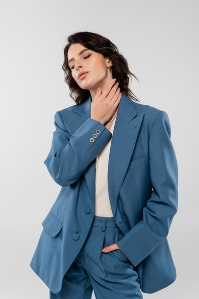 官能的なブルネット女性で青スーツ保持手でポケットと触れ首ながら立っている間に閉じられた目で隔離されたグレー - 写真・画像