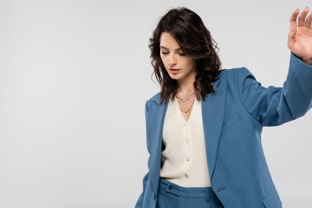 trendy brunette woman in blue blazer standing with outstretched hand isolated on grey - Fotó, kép