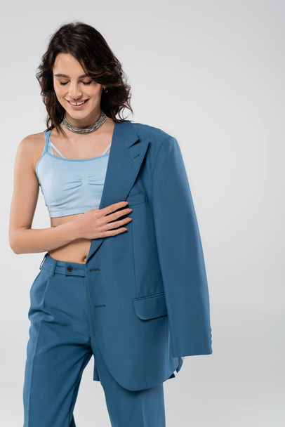 cheerful brunette woman in crop top and pants smiling with closed eyes while holding blazer isolated on grey - 写真・画像