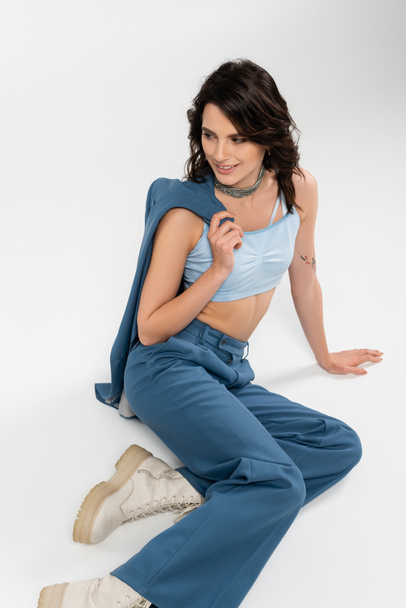 happy brunette woman in pants and boots sitting with blue jacket and looking away on grey  - Fotó, kép