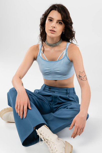 tattoed woman in blue crop top and pants with leather boots sitting with crossed legs on grey background - Фото, изображение