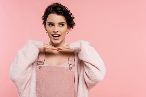 fröhliche Frau im Trägerkleid posiert mit den Händen unterm Kinn und schaut isoliert auf rosa - Foto, Bild