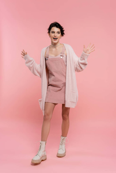 full length of excited woman in strap dress and fluffy ζακέτα που δείχνει wow χειρονομία και κοιτάζοντας μακριά σε ροζ φόντο - Φωτογραφία, εικόνα