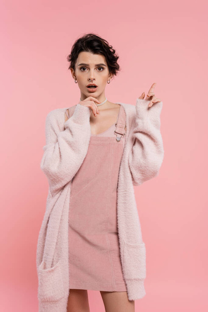 erstaunt Frau in kuscheliger Strickjacke berührt Kinn und zeigt mit dem Finger isoliert auf rosa - Foto, Bild