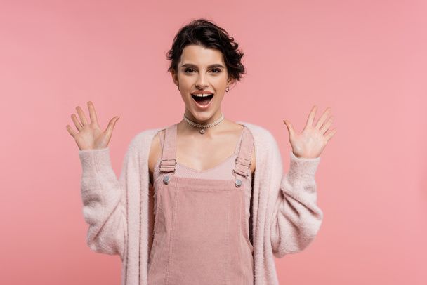 verbaasd en vrolijk vrouw in bandjurk en vest tonen wow gebaar geïsoleerd op roze - Foto, afbeelding