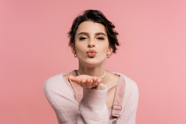 pretty brunette woman in cozy cardigan sending air kiss and looking at camera isolated on pink - Fotografie, Obrázek