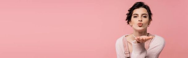 модная брюнетка женщина в теплом кардигане смотрит в камеру и отправляет воздушный поцелуй изолирован на розовый, баннер - Фото, изображение