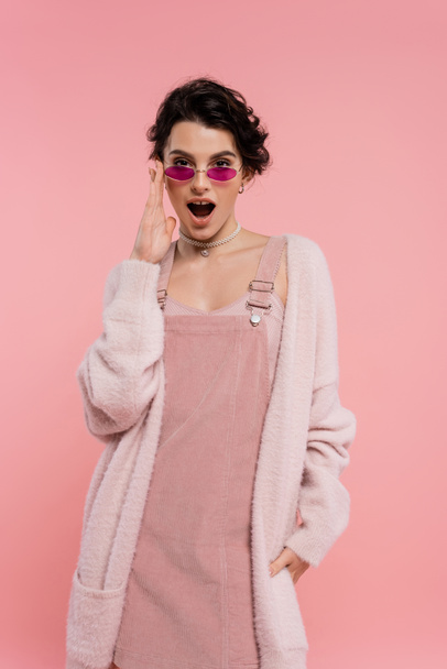 astonished woman in cozy cardigan adjusting trendy sunglasses while looking at camera isolated on pink - Photo, Image