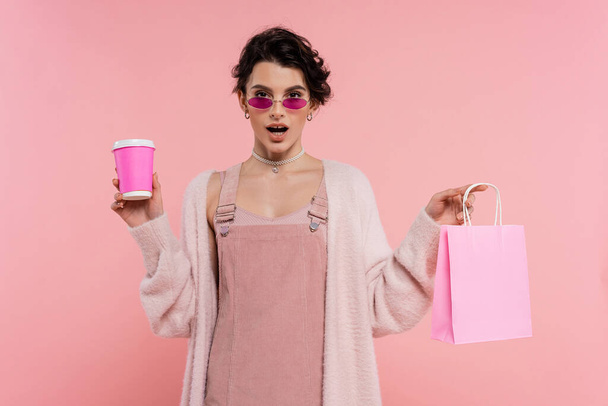 zdumiona kobieta w okularach przeciwsłonecznych i ciepłym swetrze trzymając torbę na zakupy i kubek papierowy izolowane na różowy - Zdjęcie, obraz