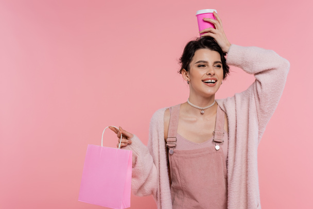 joyful and stylish woman with shopping bag holding paper cup over head isolated on pink - Zdjęcie, obraz