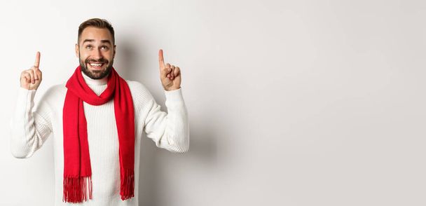 Christmas shopping and winter holidays concept. Happy bearded man in red scarf pointing fingers up, looking excited at copy space, standing over white background. - Photo, Image