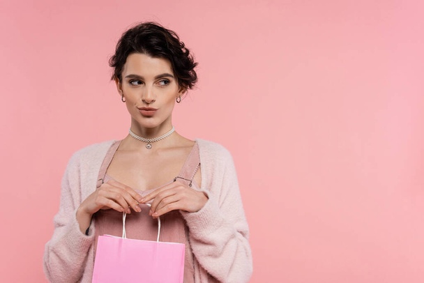 tricky brunette woman in warm cardigan holding shopping bag and looking away isolated on pink - Photo, Image