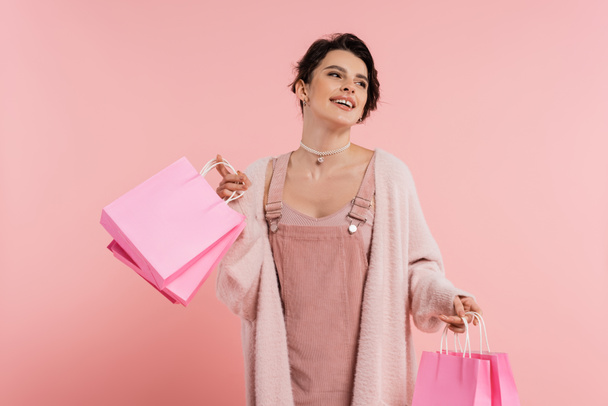 donna eccitata in abito cinturino e cardigan accogliente che tiene borse della spesa e guardando lontano isolato sul rosa - Foto, immagini