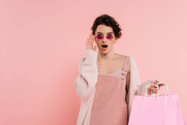 mujer asombrada con bolsas de compras tocando gafas de sol de moda y mirando a la cámara aislada en rosa - Foto, Imagen