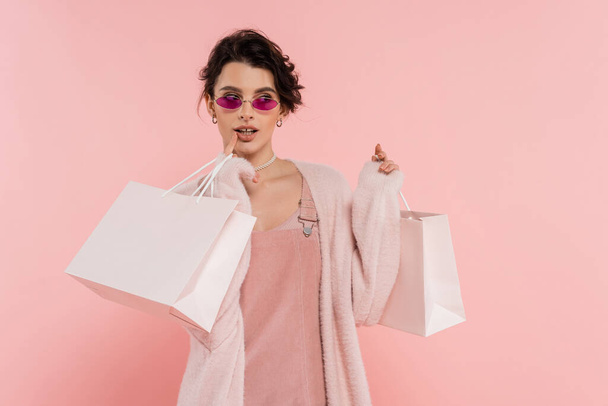 donna sognante in occhiali da sole alla moda tenendo borse della spesa e guardando lontano isolato sul rosa - Foto, immagini
