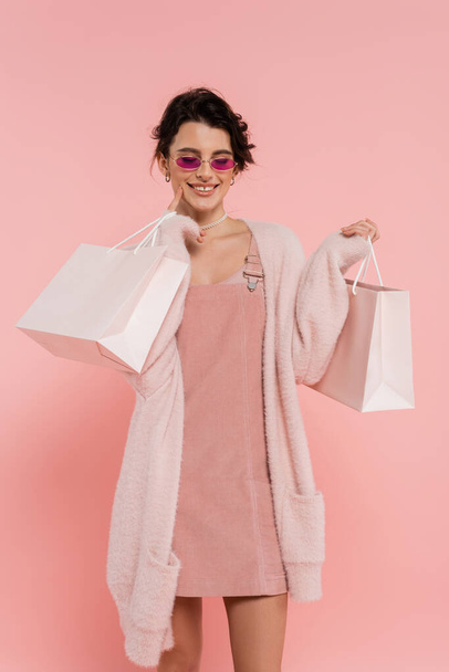 mosolygós nő divatos napszemüveg és meleg kardigán pózol bevásárló táskák elszigetelt rózsaszín - Fotó, kép