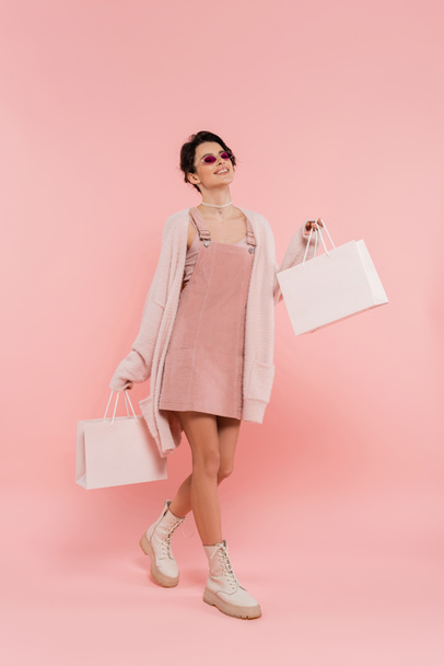 donna sorridente in occhiali da sole e abbigliamento alla moda che cammina con borse della spesa su rosa  - Foto, immagini