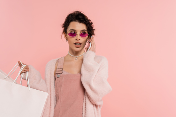 donna bruna in occhiali da sole alla moda che tiene le borse della spesa e parla su smartphone isolato su rosa - Foto, immagini