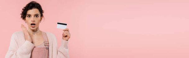 zdumiona kobieta w ciepłym swetrze z kartą kredytową i patrząc na aparat odizolowany na różowy, baner - Zdjęcie, obraz