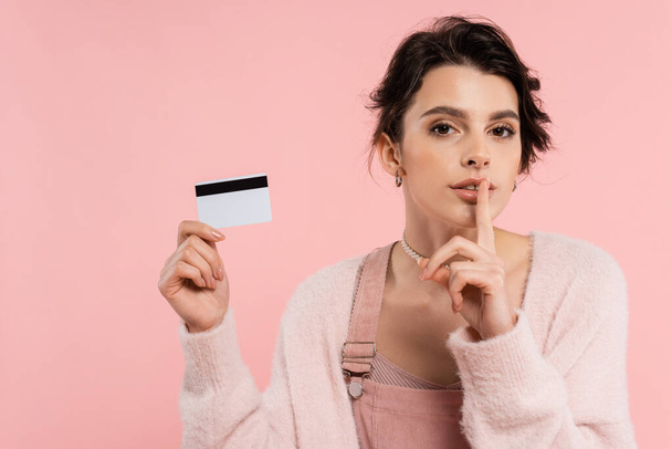 mladá brunetka žena v útulný svetr ukazující ticho znamení při držení kreditní karty izolované na růžové - Fotografie, Obrázek