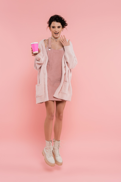 full length of excited woman in strap dress and cozy cardigan levitating with paper cup on pink background - Фото, изображение