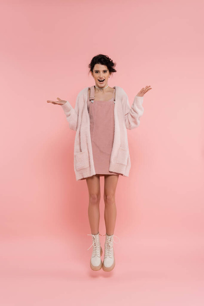 full length of excited woman in strap dress and fluffy cardigan levitating on pink background - Photo, Image