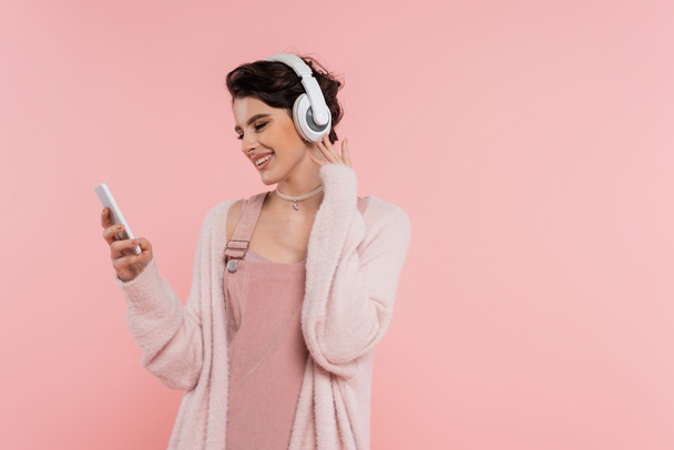 mujer morena feliz en auriculares inalámbricos mirando el teléfono móvil aislado en rosa - Foto, Imagen
