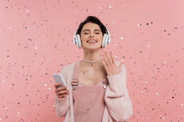 radostná žena se zavřenýma očima držící mobilní telefon a poslouchající hudbu v bezdrátových sluchátkách poblíž konfety na růžové  - Fotografie, Obrázek