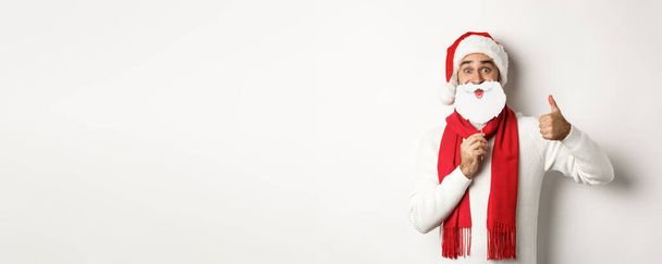 Weihnachtsfeier und Festkonzept. Glückliches männliches Model mit Nikolausmütze und weißer Bartmaske, Daumenhoch-Geste, vor weißem Hintergrund stehend. - Foto, Bild