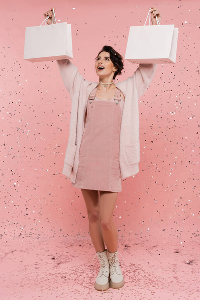 full length of excited and fashionable woman holding shopping bags in raised hands under festive confetti on pink background - Photo, Image