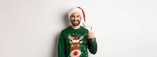 Christmas party and holidays concept. Happy man in Santa hat and sweater smiling, making a toast with glass of champagne, standing over white background. - Photo, Image