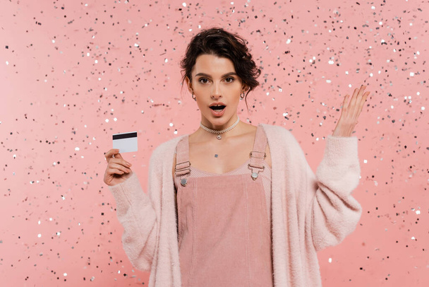 mulher espantada em vestido de cinta e casaco de lã segurando cartão de crédito e apontando com a mão sob confete no fundo rosa - Foto, Imagem
