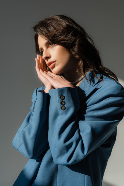 thoughtful brunette woman in blue blazer standing with hands near face and looking away on grey  - Foto, imagen