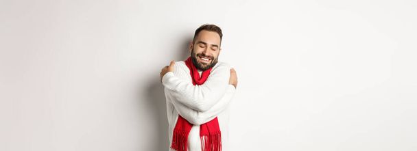 Happy bearded man enjoying comfortable winter sweater, smiling with warmth, hugging himself, standing over white background. - Fotó, kép