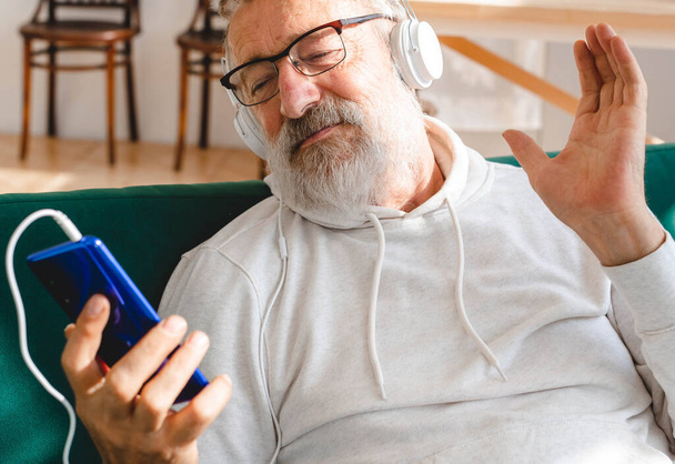 Idős ember hűvös szakállas öregember használja a mobiltelefon videohívás - boldogság idős életmód emberek koncepció. Videohívás okostelefonon a szobában integetett a képernyőn, és beszélgetni a gyerekekkel - modern - Fotó, kép
