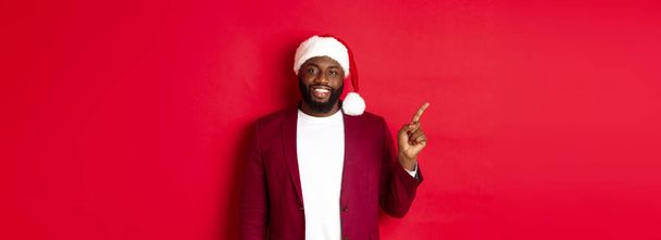 Koncept Vánoc, večírků a prázdnin. Fešák černoch v Santa klobouku ukazující prstem vlevo, ukazující reklamu, stojí šťastně nad červeným pozadím. - Fotografie, Obrázek