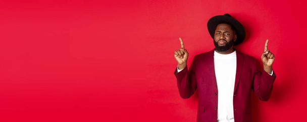 Winter holidays and shopping concept. Skeptical Black man grimasing and showing dislike, pointing fingers up at bad offer, standing over red background. - Photo, Image