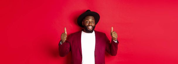 Divat és party koncepció. Vidám afro-amerikai férfi elegáns kabát, ünnepli újévi karácsony, mutatja hüvelykujját fel és mosolyog, mint és jóváhagyja, piros háttér. - Fotó, kép