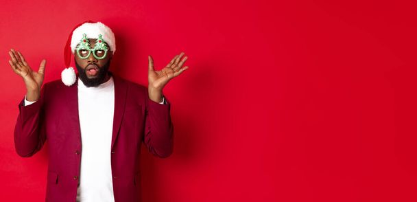 Merry Christmas. Cheerful Black man wearing funny party glasses and santa hat, smiling joyful, celebrating winter holidays, standing over red background. - Foto, Imagem