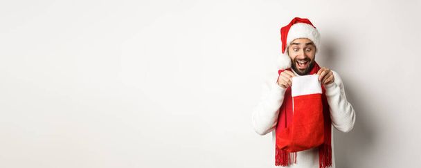 Zimowe wakacje i koncepcja świętowania. Szczęśliwy facet patrząc wewnątrz skarpetki świąteczne, otrzymując prezent noworoczny, stojąc nad białym tle. - Zdjęcie, obraz